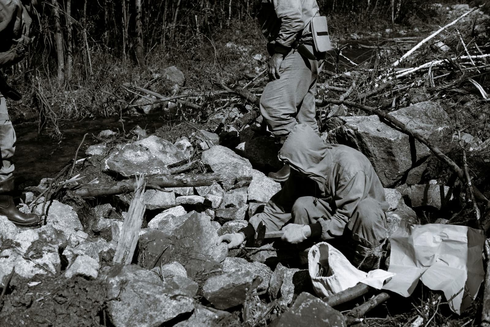 Two field geologists in hooded jackets examining broken rock beside a forest stream, faces turned away, in black and white, under the headline Grassroots to Resource