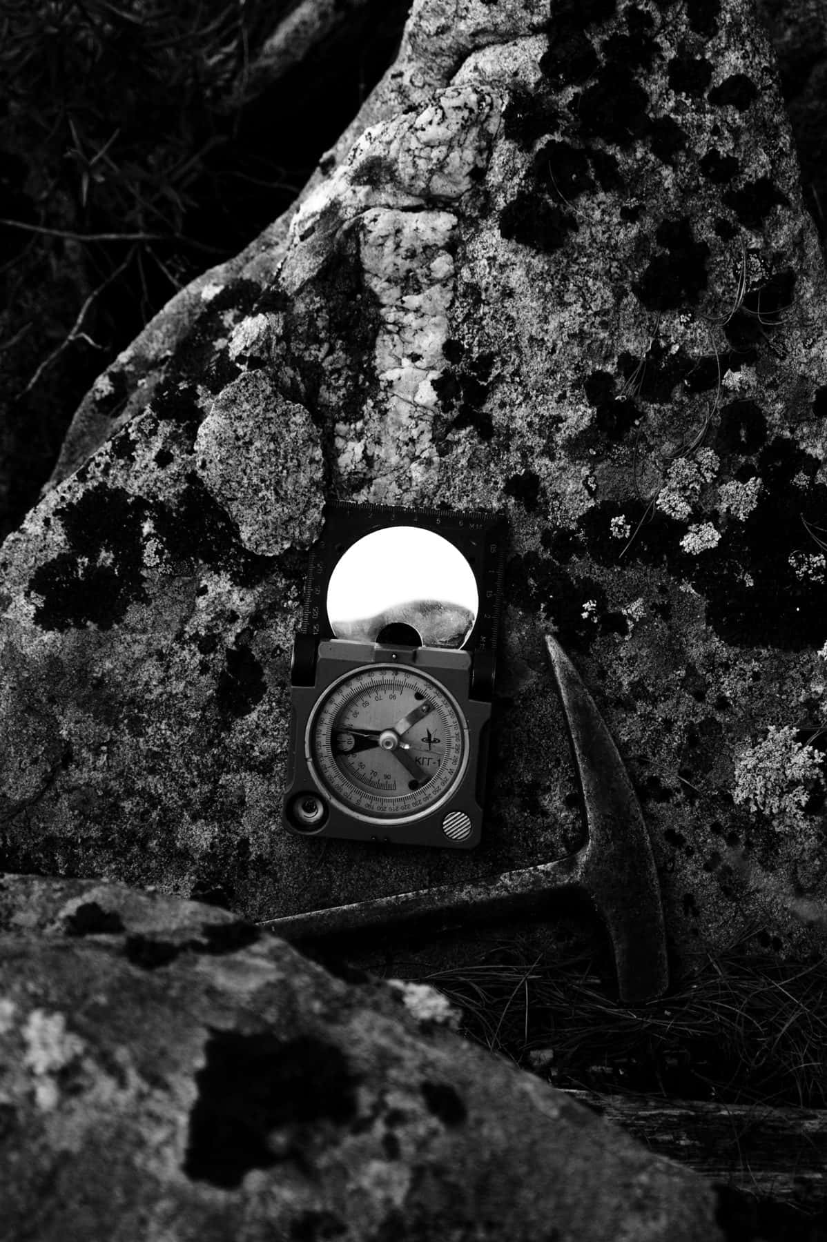 A geological compass and rock hammer resting on a lichen-covered boulder, in black and white, under the headline Finding Targets