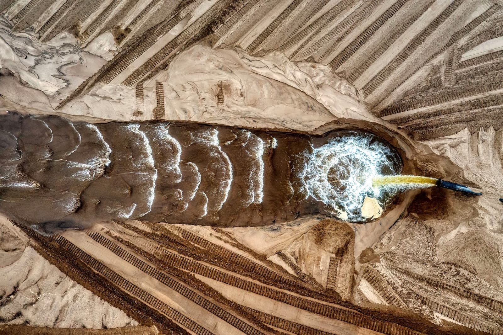 Aerial view of grey slurry flowing from a pipe into a sandy basin, under the headline Tailings